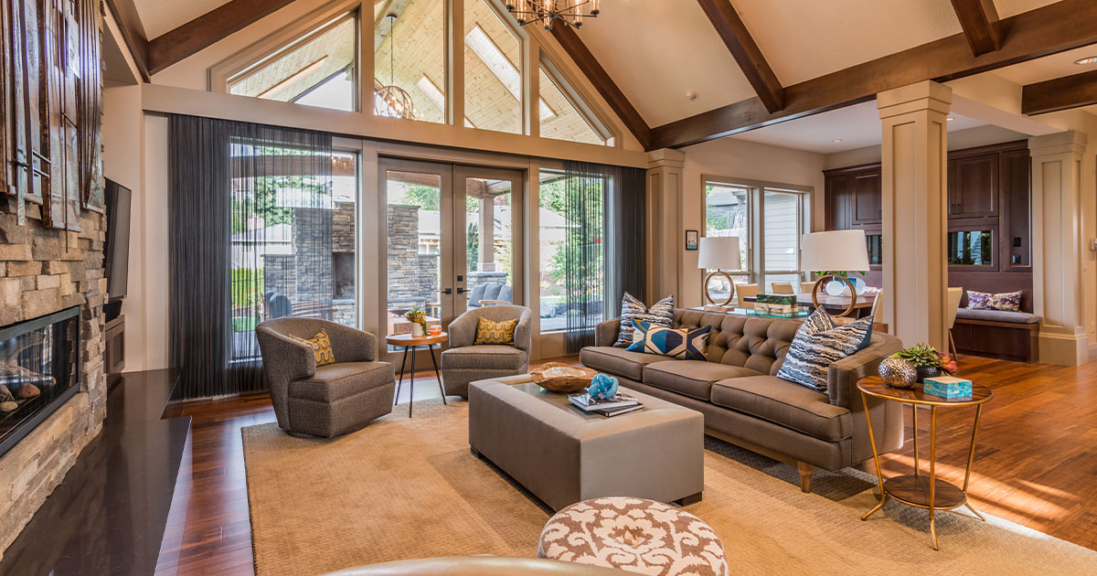 Elegant living room with vaulted ceiling, exposed wooden beams, cozy stone fireplace, and large windows for ample natural light, perfect for luxurious home remodeling.