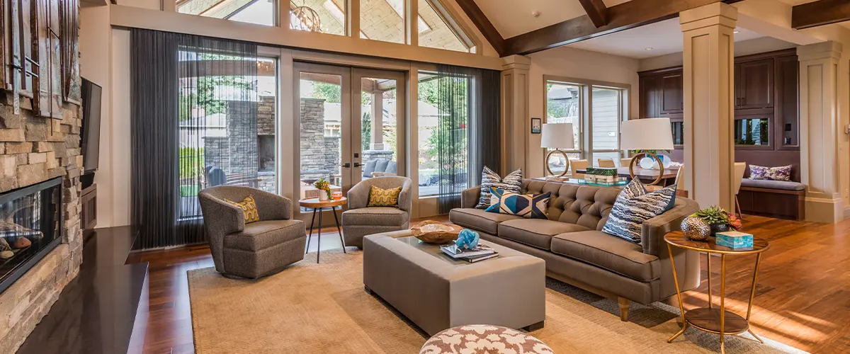 Elegant living room with vaulted ceiling, exposed wooden beams, cozy stone fireplace, and large windows for ample natural light, perfect for luxurious home remodeling.