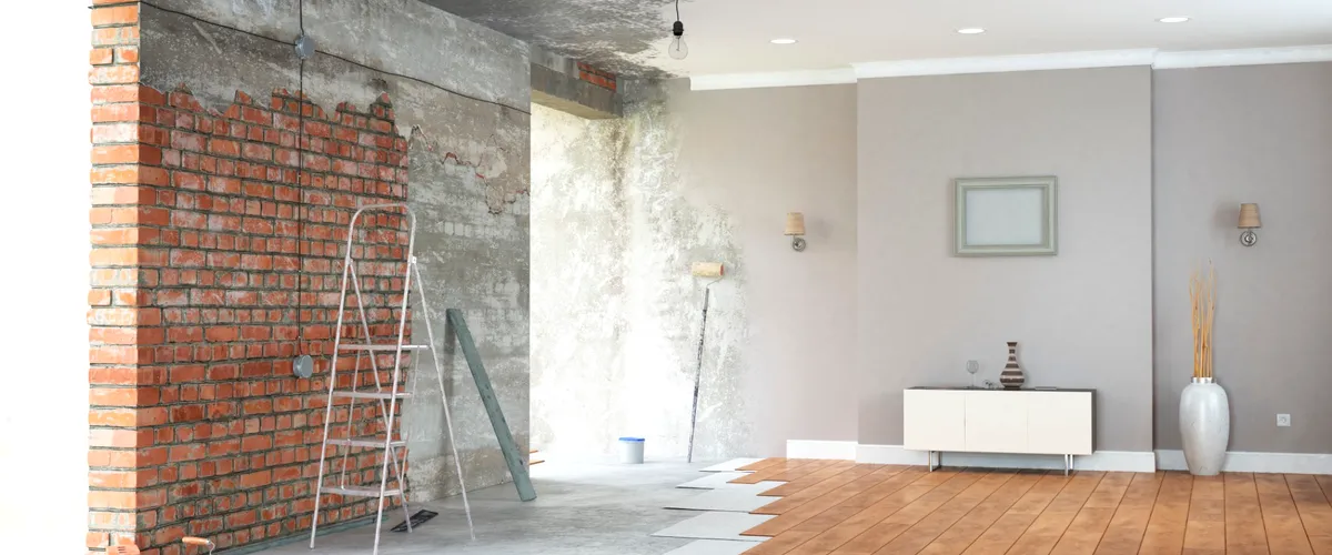 Interior room showing renovation progress with exposed brick wall, partially painted surfaces, and wooden flooring installation