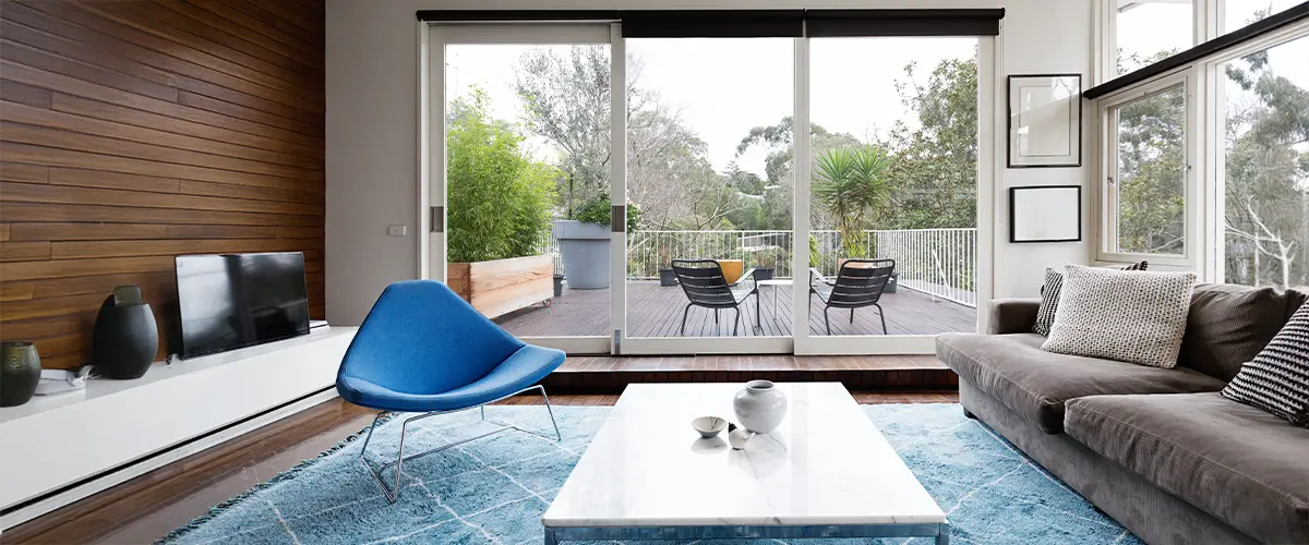 Modern home addition featuring a glass sliding door that opens to a deck with seating, surrounded by greenery.