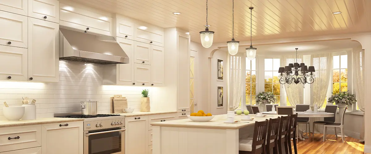 Spacious white kitchen with island seating, pendant lights, and a dining area featuring a chandelier and large windows.