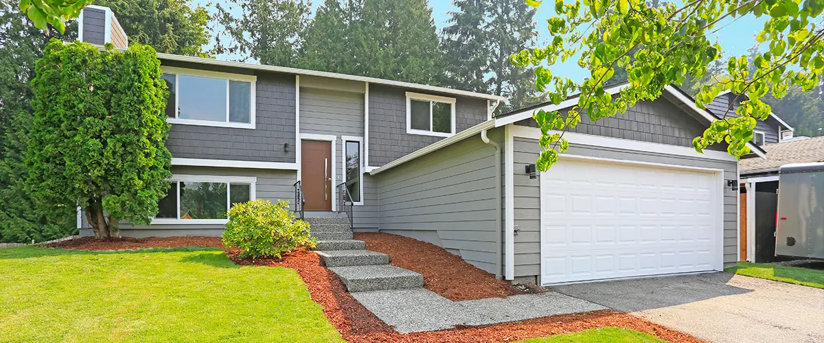 Modern gray split-level home with landscaped yard, white garage, and concrete walkway.