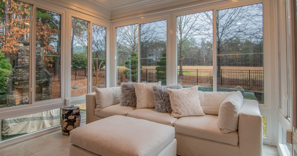 Cozy sunroom with large windows, a plush sectional sofa, and an outdoor fireplace in view.
