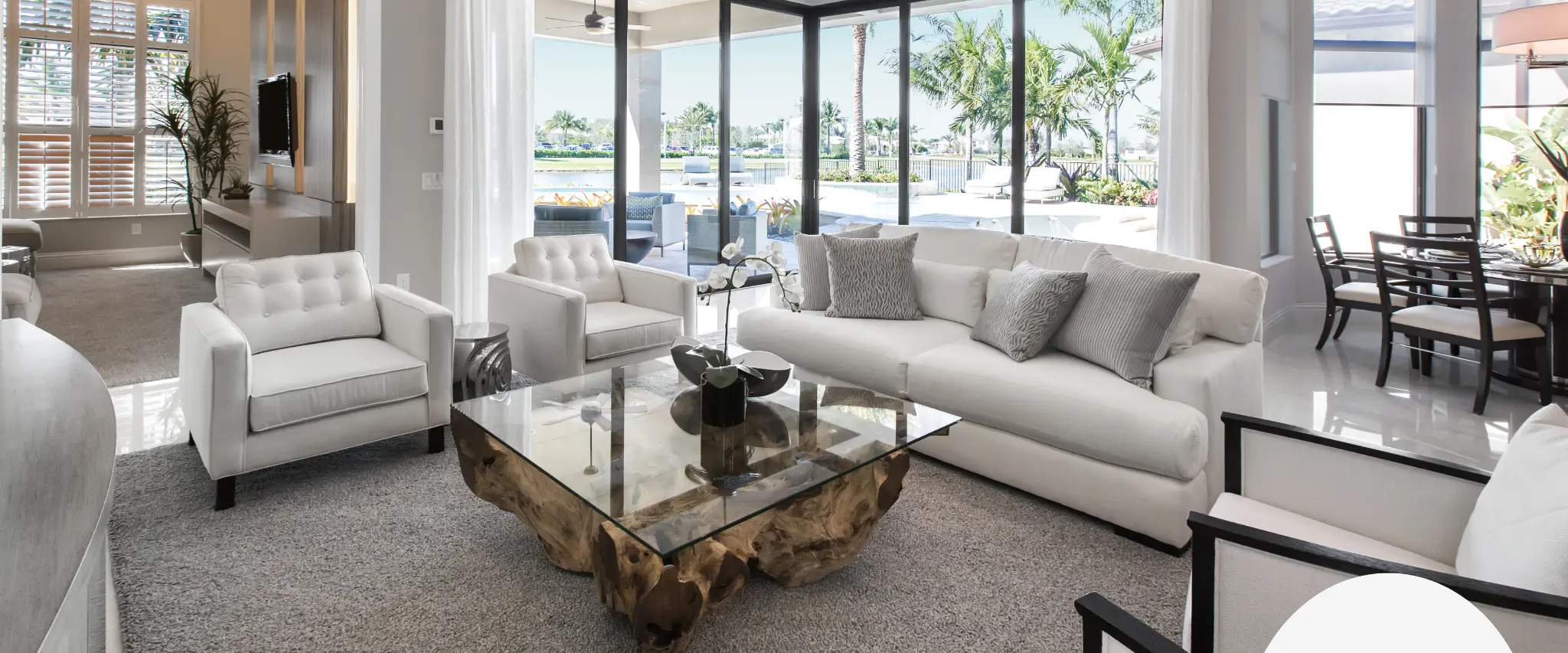 A bright and modern living room with a glass coffee table and cozy white sofas, overlooking a tropical outdoor area.