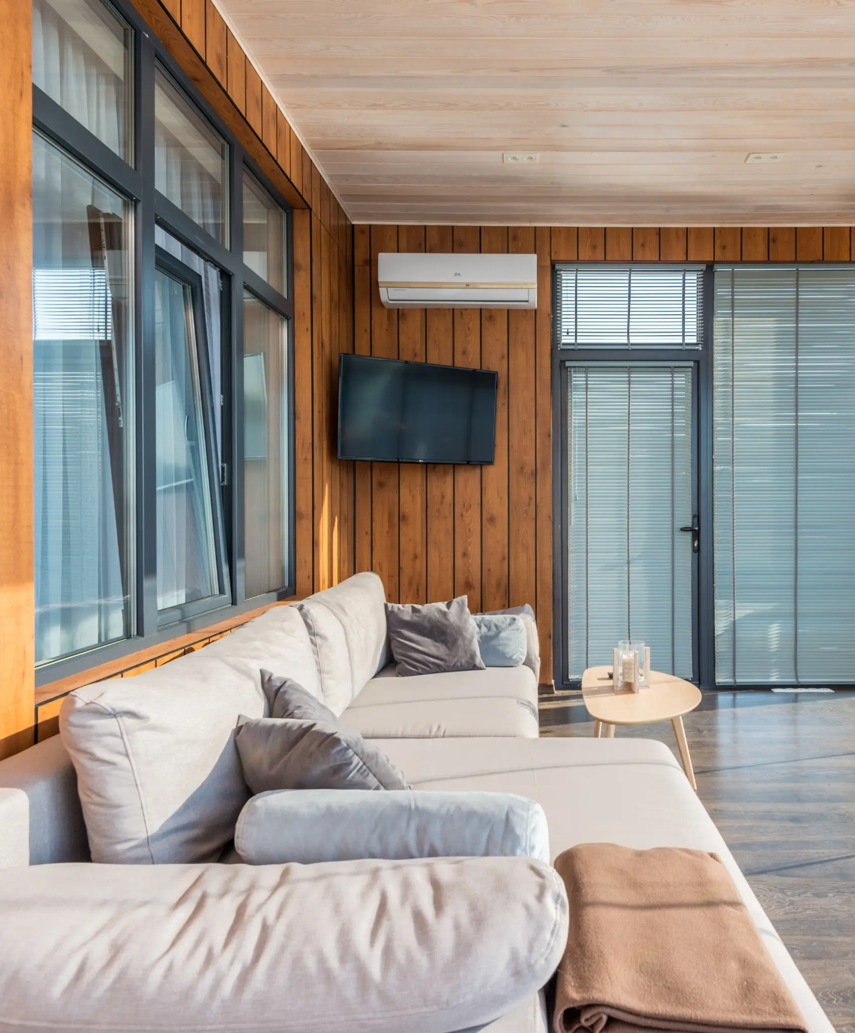 A rustic-style sunroom with warm wood-paneled walls, large windows, and a comfortable light gray sectional sofa. A mounted TV and modern décor complete the inviting atmosphere.