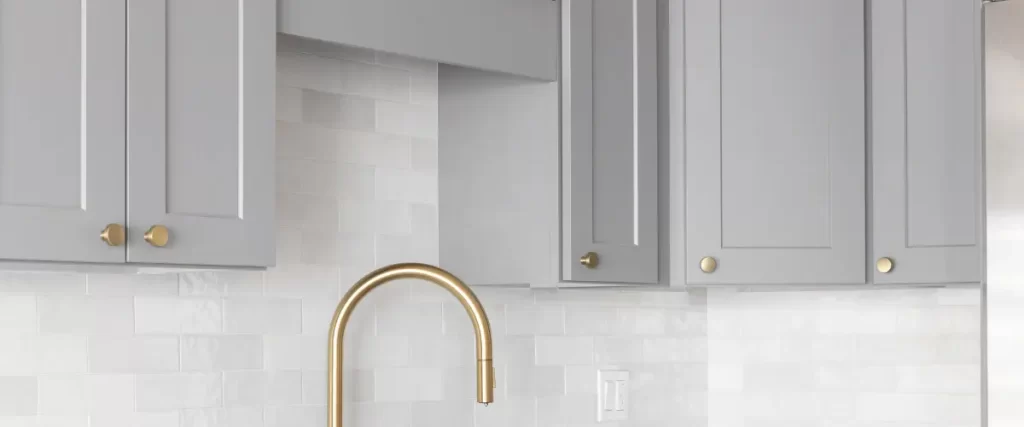 A gold kitchen faucet mounted on a white subway tile backsplash beneath gray cabinets.