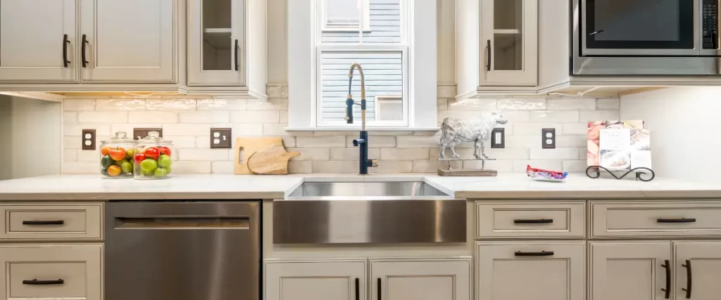 A farmhouse-style kitchen sink with a high-arc faucet, surrounded by beige cabinets and a marble backsplash.