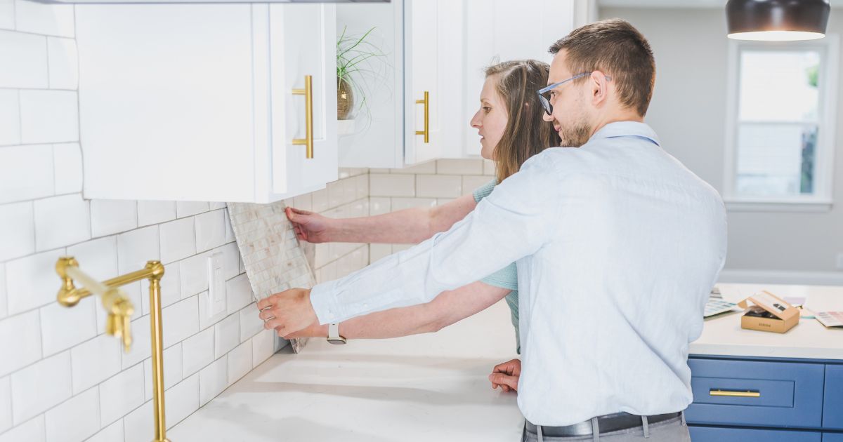 A couple installing a white subway tile backsplash in a kitchen, aligning the tiles carefully.