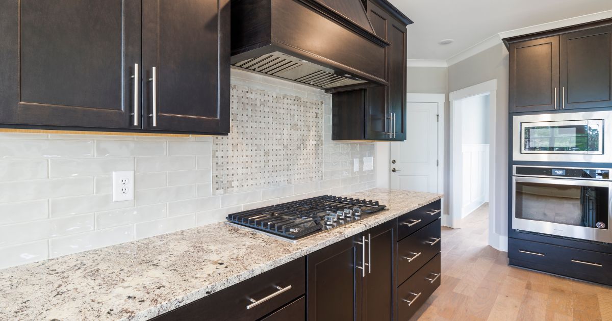 A sleek kitchen with dark cabinetry, a mosaic tile backsplash, and a beige granite countertop with a built-in cooktop.