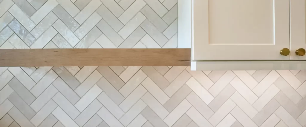 A close-up of a herringbone-patterned tile backsplash with a wooden shelf above it.