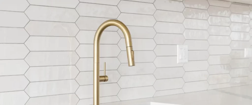 A gold kitchen faucet mounted on a white hexagonal tile backsplash.